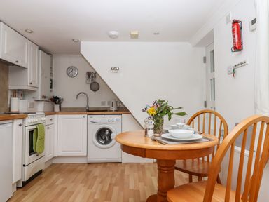 A kitchen with white cabinets wooden counter a washing machine a round wooden dining table with two chairs and tableware at Wisteria Cottage in Exmouth