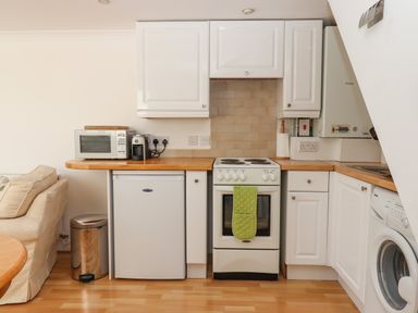 A kitchen with white cabinets a microwave a coffee machine a small fridge an oven with a green glove a washing machine and a silver trash can at Wisteria Cottage in Exmouth