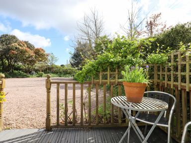 A small outdoor wooden deck with a metal table and chair and a potted plant with greenery and trees in the background at Wisteria Cottage in Exmouth