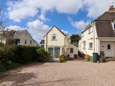 A gravel driveway with hedges and two cream houses under a blue sky at Wisteria Cottage in Exmouth