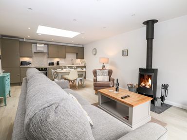 An open-plan living room and kitchen with a grey sofa a wood stove a dining table and kitchen cabinets at Rabbit's Warren in Titchfield Common