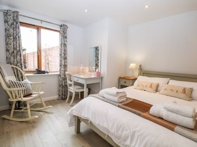A bedroom with a bed, rocking chair, dressing table with mirror, bedside table, and window with curtains at Rabbits Warren in Titchfield Common