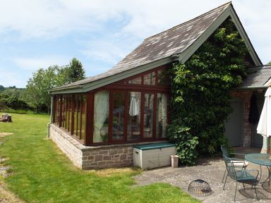 A glass conservatory attached to a stone house with climbing plants and outdoor furniture on a patio at The Granary in Walford near Ross-on-Wye