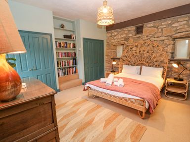 A bedroom with a double bed with rolled towels on a pink blanket a wooden dresser a woven rug bookshelf built into the wall and stone wall behind the bed at Barn Cottage in Marazion