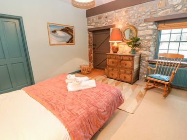 A bedroom with a bed with folded towels a wooden dresser with a lamp and plant a rocking chair and a window at Barn Cottage in Marazion