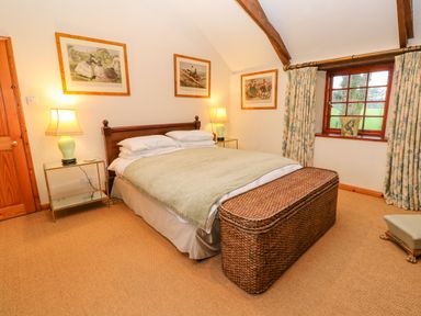 A bedroom with a double bed bedside tables with lamps wicker chest at the foot of the bed window with curtains and paintings on the walls at Barn Cottage in Marazion