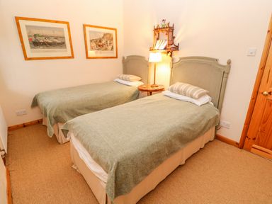 A bedroom with two single beds a round wooden side table and framed pictures on the wall at Barn Cottage in Marazion