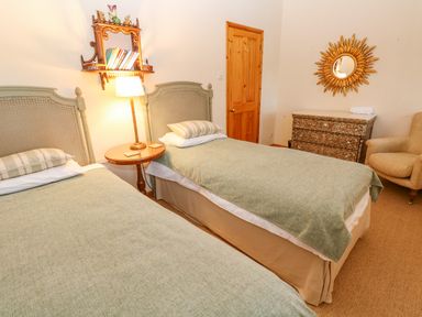 A bedroom with two single beds a wooden side table with a lamp a wall shelf with books a wooden door a dresser with a sunburst mirror and an armchair at Barn Cottage in Marazion