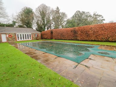 A covered swimming pool with stone patio and green grass near a stone pool house with white doors and windows at Barn Cottage in Marazion