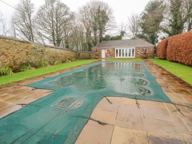 An outdoor pool covered with a green safety cover surrounded by stone paving grass and trees at Barn Cottage in Marazion