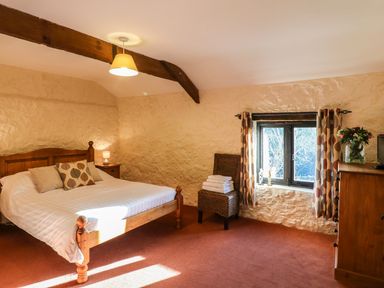 A bedroom with a wooden bed and nightstand next to a window with curtains and a wicker chair at Carthorse Cottage in Teignmouth