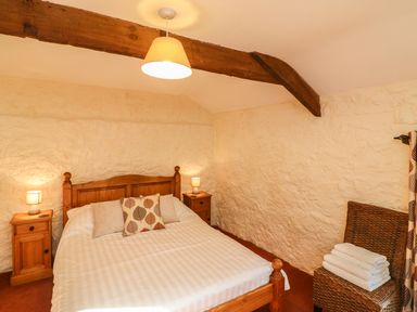A bedroom with a wooden bed with three pillows two bedside tables with lamps a wicker chair with folded towels and exposed ceiling beam at Carthorse Cottage in Teignmouth