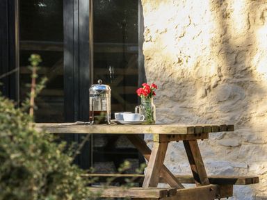 A wooden picnic table with a glass coffee press a white cup and saucer and a jar of red flowers outside by a stone wall