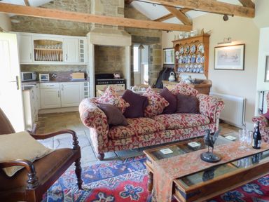 A living room with a patterned sofa and armchairs a glass coffee table and a kitchen with white cabinets at Humbleton Cottage Haydon Bridge near Hexham