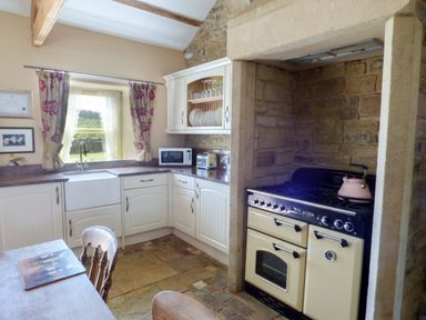 A kitchen with white cabinets a farmhouse sink a stove with a pink kettle and curtains on a window at Humbleton Cottage in Haydon Bridge near Hexham