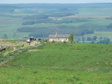 A stone cottage on a green hill with vehicles and outbuildings surrounded by fields and distant hills at Humbleton Cottage in Haydon Bridge near Hexham