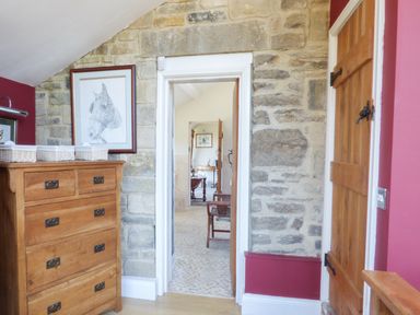 A hallway with stone walls and a wooden chest of drawers beside a wooden door leading to a room with wooden chairs at Humbleton Cottage in Haydon Bridge near Hexham