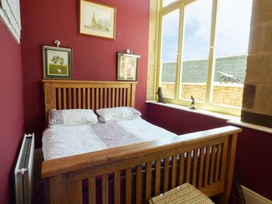 A bedroom with a wooden bed frame red walls three paintings and a large window at Humbleton Cottage in Haydon Bridge near Hexham