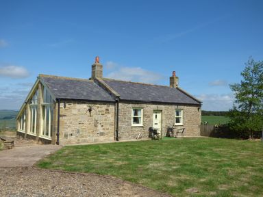 A stone cottage with two chimneys a small patio with table and chairs and surrounding grass at Humbleton Cottage in Haydon Bridge near Hexham