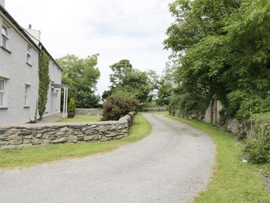 A gravel driveway curves past a white house with stone walls and greenery at Bodegri Cottage in Llanrhyddlad near Church Bay
