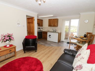 A living room with kitchen area a round dining table and chairs a black armchair sofa with cushions a red rug and a wooden side table with flowers at Bodegri Cottage Llanrhyddlad near Church Bay