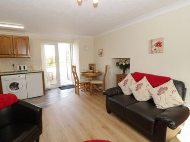 A living room with brown sofa and armchair a small dining table and kitchenette with washing machine and fridge at Bodegri Cottage in Llanrhyddlad near Church Bay