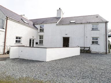 An exterior view of a white house with multiple windows a chimney two skylights a gravel yard and a low white wall at Bodegri Cottage in Llanrhyddlad near Church Bay
