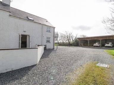 Exterior view of a white two-story house with open doors and gravel driveway with covered parking and cars at Bodegri Cottage Llanrhyddlad near Church Bay
