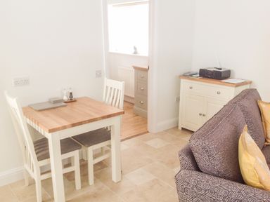 A dining area with a wooden table and two chairs next to a grey patterned sofa and a cabinet with a radio at The Hideaway in Verwood