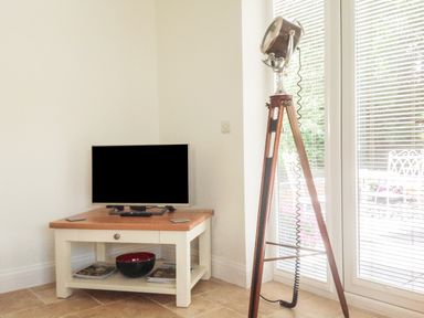 A television on a wooden table next to a floor lamp in front of a window with blinds at The Hideaway in Verwood