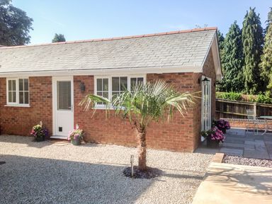 A small brick building with a white door and windows surrounded by gravel and a palm tree at The Hideaway in Verwood