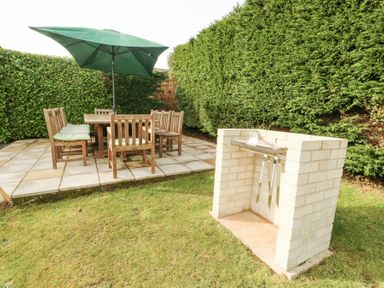 An outdoor patio area with a wooden table and chairs under a green umbrella and a built-in brick barbecue grill in a garden at Bay View in Benllech