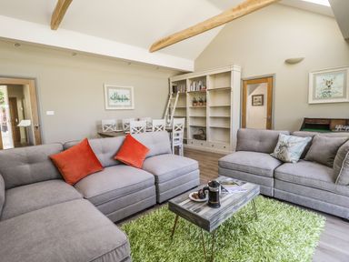 A living room with gray sofas and orange and patterned cushions a wooden coffee table on a green rug and a dining table with white chairs and bookshelves at Providence Cottage in Easingwold