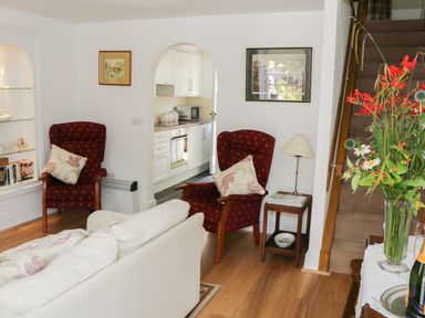 A living room with two red armchairs a white sofa a wooden side table a lamp flowers on a table and stairs with a kitchen visible through an archway at Coachmans Cottage in Witherslack
