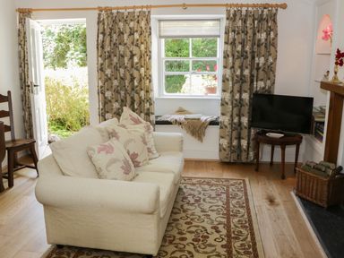 A living room with a beige sofa and floral cushions a patterned rug curtains on windows a television on a wooden table and a basket with logs at Coachmans Cottage in Witherslack