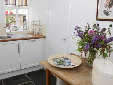 A kitchen corner with a wooden countertop sink and cabinets a table with a flower vase and a bread box at Coachmans Cottage in Witherslack