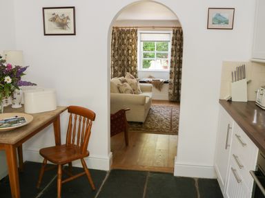 A kitchen area with wooden chair and table next to a living room with sofa and window at Coachmans Cottage in Witherslack