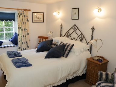 A bedroom with a double bed with pillows and folded towels a wooden nightstand with a lamp and a window seat with cushions at Coachmans Cottage in Witherslack