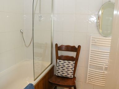 A bathroom with a bathtub that has a glass shower screen a wooden chair with a cushion a round mirror and a white towel radiator at Coachmans Cottage in Witherslack