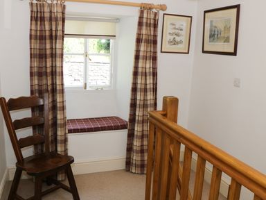 A corner with a wooden chair next to a window with checkered curtains and cushion and a wooden railing in Coachmans Cottage in Witherslack