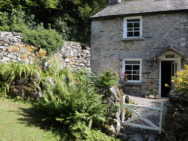Exterior of a stone cottage with white framed windows a small white gate and a garden with flowers and plants at Coachmans Cottage in Witherslack