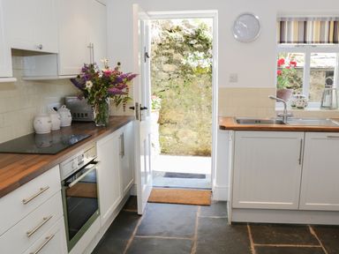 A kitchen with white cabinets wooden countertops a sink a clock on the wall and a door open to a stone wall outside at Coachmans Cottage in Witherslack