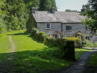 A stone cottage with chimneys surrounded by grass and trees at Coachmans Cottage in Witherslack