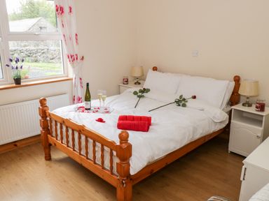A bedroom with a wooden bed with white bedding two red roses and red towels on the bed a bottle of wine with two glasses on a tray next to the window with curtains and two bedside tables with lamps and candles at Lough Mask Road Fishing Cottage in Ballinrobe County Mayo