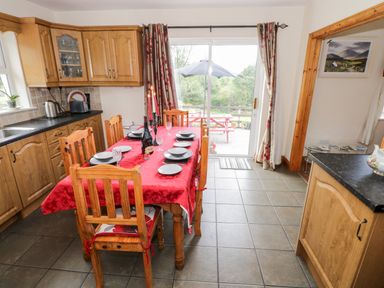 A kitchen with wooden cabinets a dining table with red tablecloth and plates and chairs near a sliding glass door at Lough Mask Road Fishing Cottage in Ballinrobe County Mayo