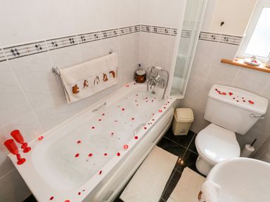 A bathroom with a bathtub filled with bubbles and rose petals a toilet with rose petals on the tank a towel rack with a towel featuring horse designs and a window with items on the sill at Lough Mask Road Fishing Cottage in Ballinrobe County Mayo