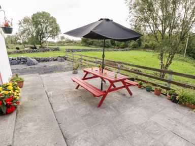 An outdoor patio with a red picnic table and black umbrella surrounded by potted plants and green fields at Lough Mask Road Fishing Cottage Ballinrobe County Mayo