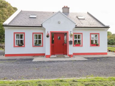 A white house with a red door and window frames surrounded by greenery at Lough Mask Road Fishing Cottage in Ballinrobe County Mayo