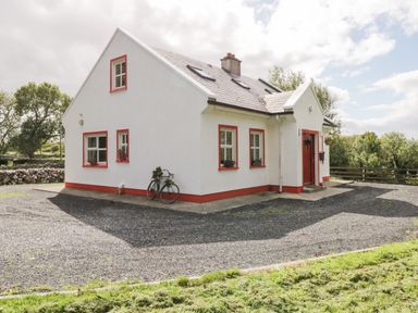 A white house with red trim around windows and door a bicycle leaning against the wall and trees in the background at Lough Mask Road Fishing Cottage in Ballinrobe County Mayo