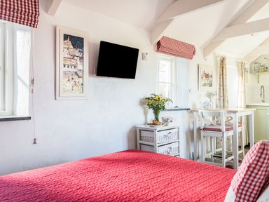 A bedroom with a red bedspread a wall-mounted TV a small table with flowers and a dining area at The Snug in Rescorla near St Austell